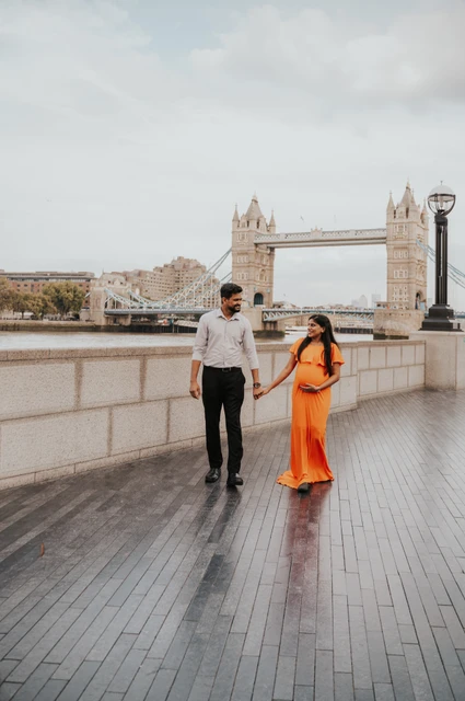 London maternity photoshoot: a wide promenade view of Tower Bridge. A couple hold hands on the Queen’s Walk promenade, with the woman’s long orange dress and Tower Bridge visible in the wide riverside setting during their London maternity photoshoot.