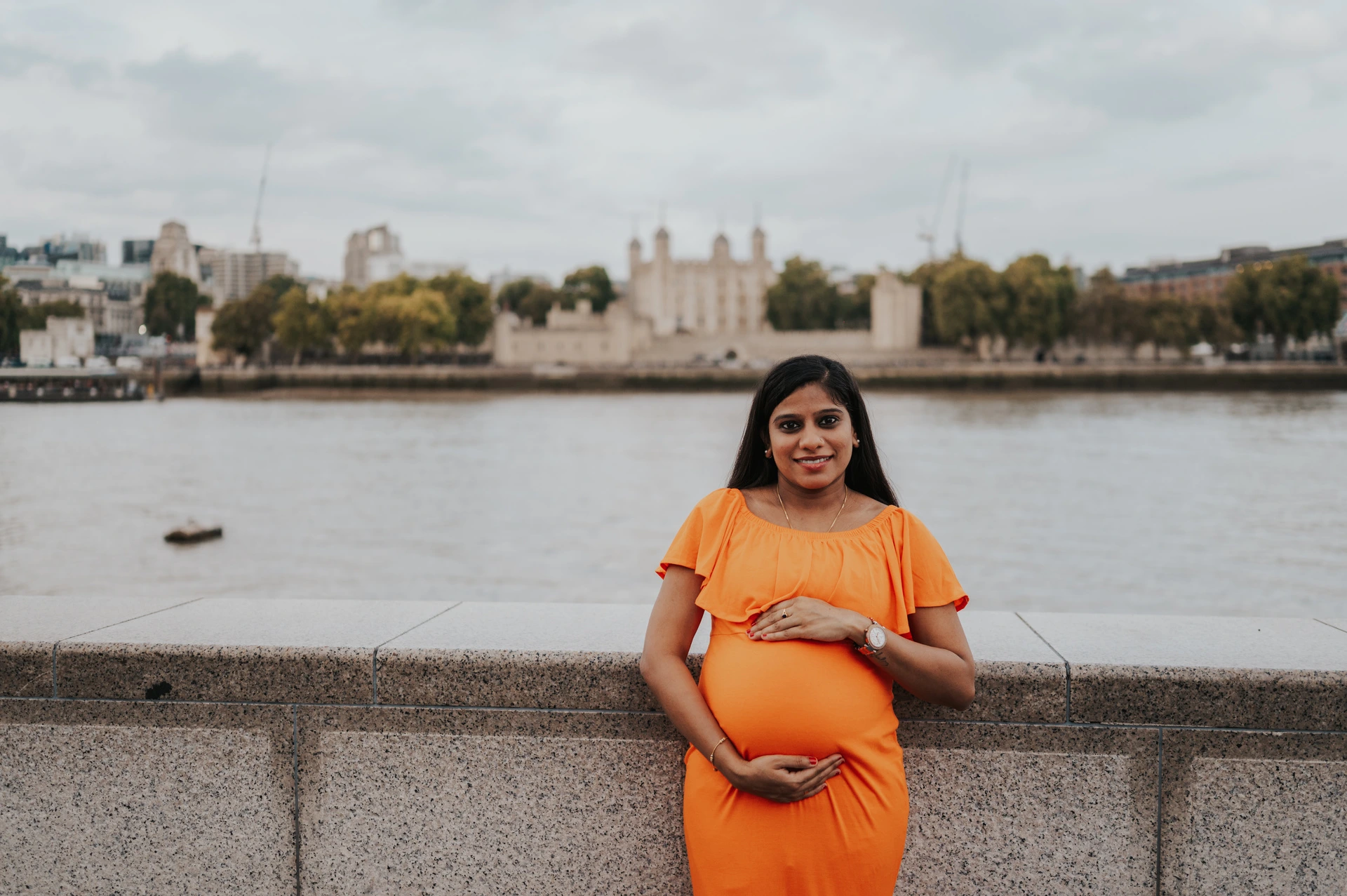 A wide portrait shows a pregnant woman holding her bump in an orange dress beside the stone wall, with the Thames and Tower of London behind during their London maternity photoshoot.