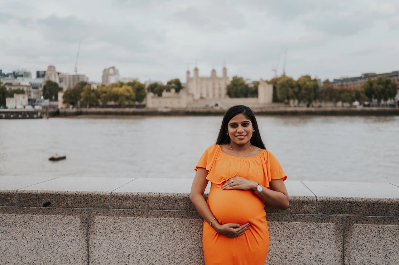 London maternity photoshoot: a wide maternity portrait across from the Tower of London. A wide portrait shows a pregnant woman holding her bump in an orange dress beside the stone wall, with the Thames and Tower of London behind during their London maternity photoshoot.