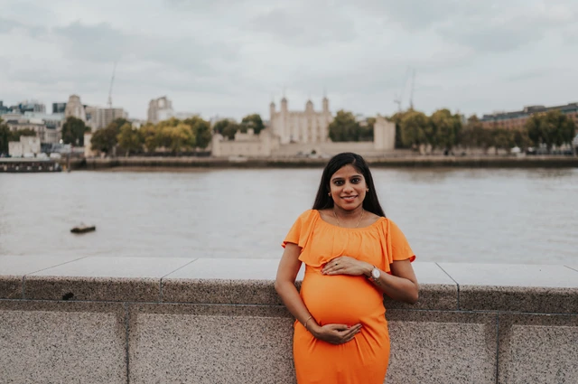 A wide portrait shows a pregnant woman holding her bump in an orange dress beside the stone wall, with the Thames and Tower of London behind during their London maternity photoshoot.