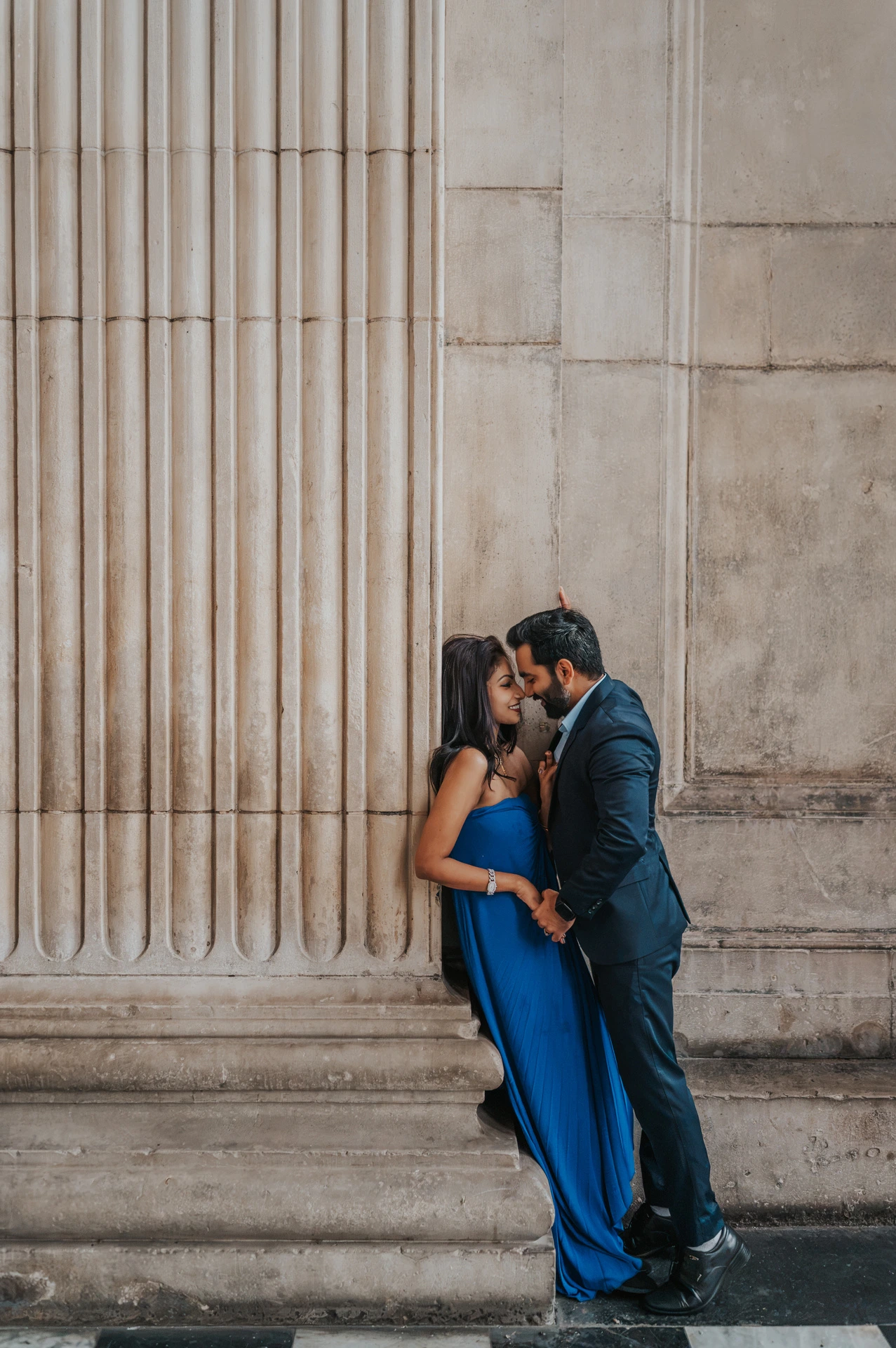 The couple leaning close together beside a fluted stone column at St Paul’s Cathedral during their London proposal photoshoot.