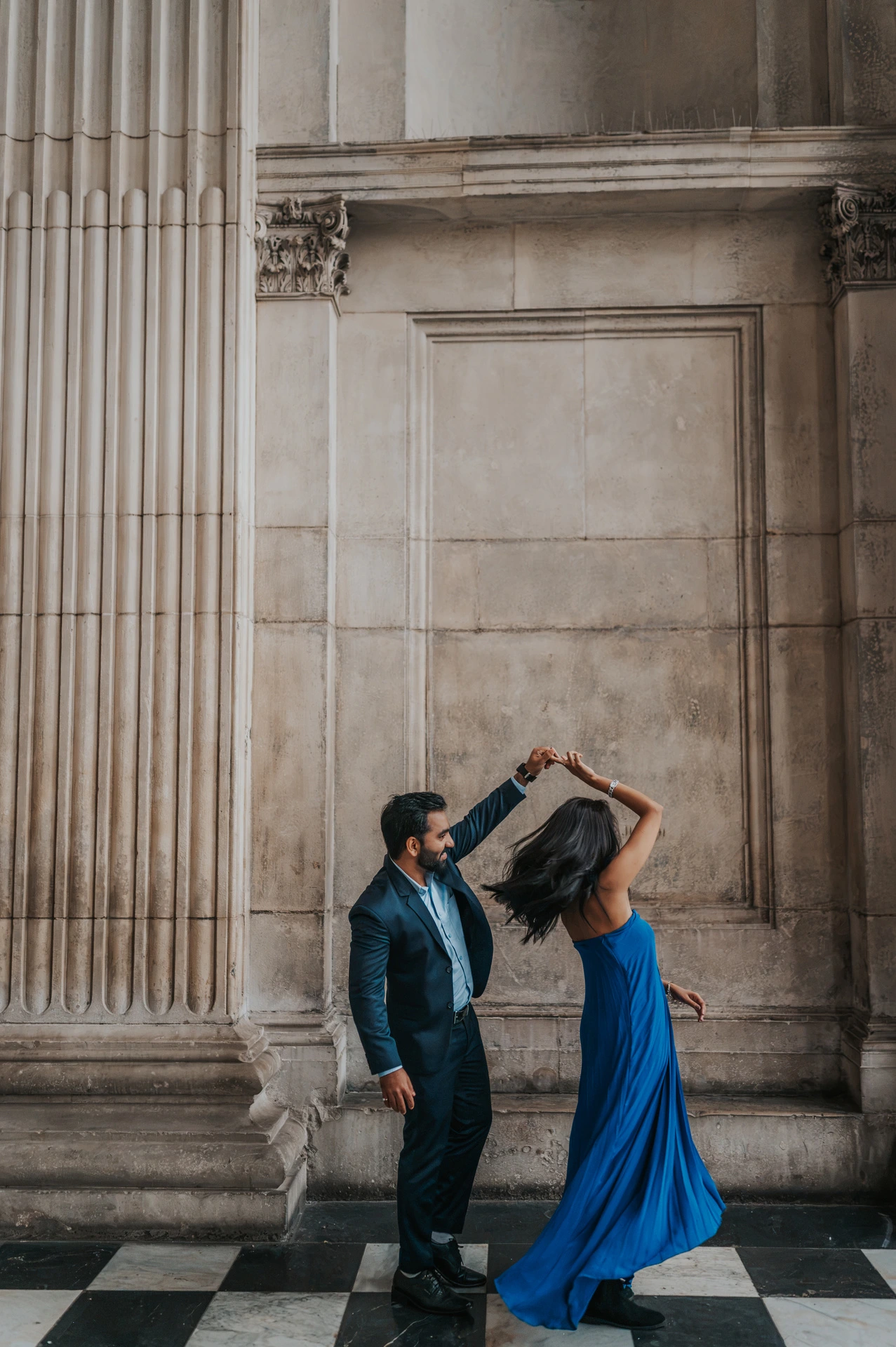 The man lifting his partner’s hand as she twirls in a blue dress between the columns of St Paul’s Cathedral during their London proposal photoshoot.