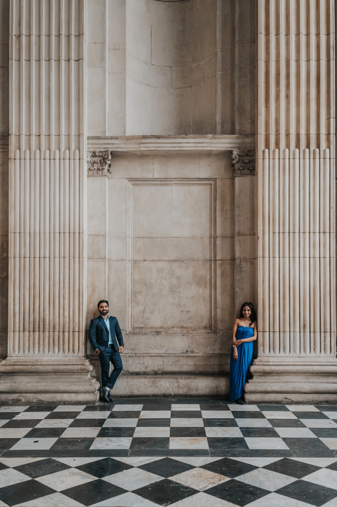 The couple standing apart between the tall columns of St Paul’s Cathedral above a checkerboard floor during their London proposal photoshoot.