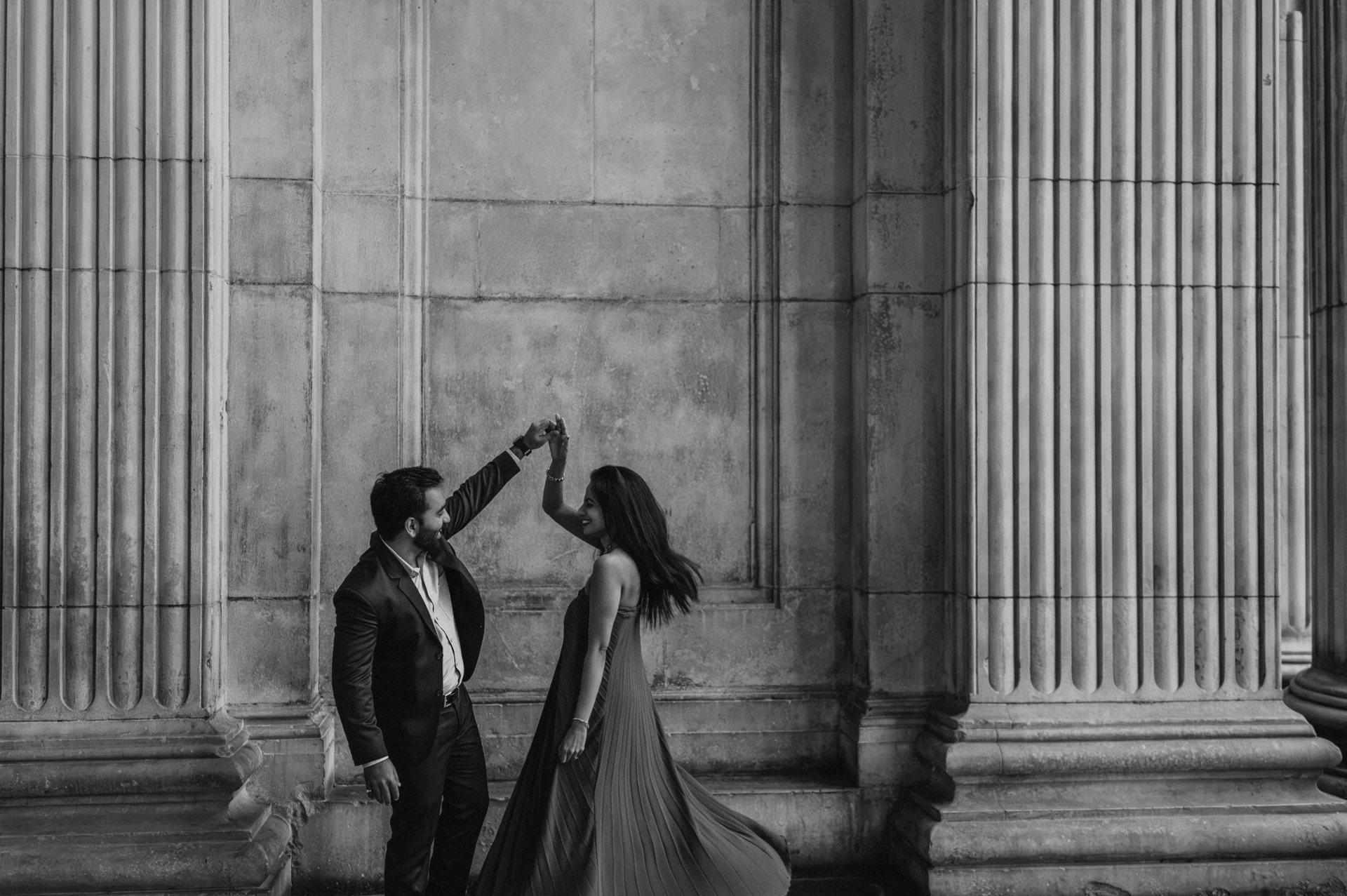 Black-and-white photograph of a wide view of the couple with their joined hands raised between the columns of St Paul’s Cathedral during their London proposal photoshoot.