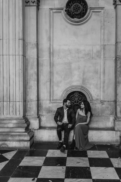 London proposal photoshoot: a seated cathedral portrait in black and white Black-and-white photograph of the couple sitting together against the cathedral wall above the checkerboard floor at St Paul’s Cathedral during their London proposal photoshoot.