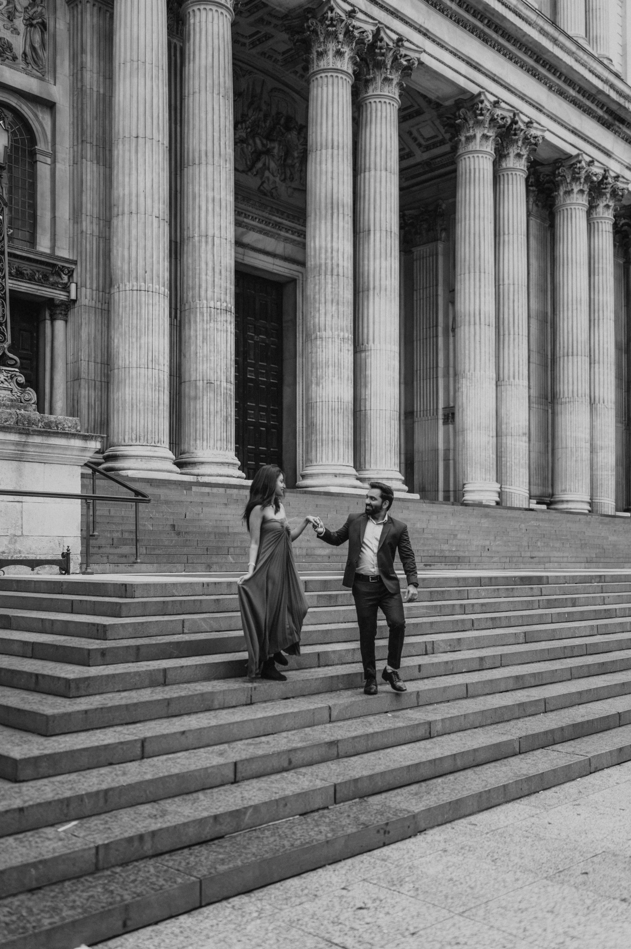 Black-and-white photograph of the couple walking hand in hand on the broad steps below the columns of St Paul’s Cathedral during their London proposal photoshoot.