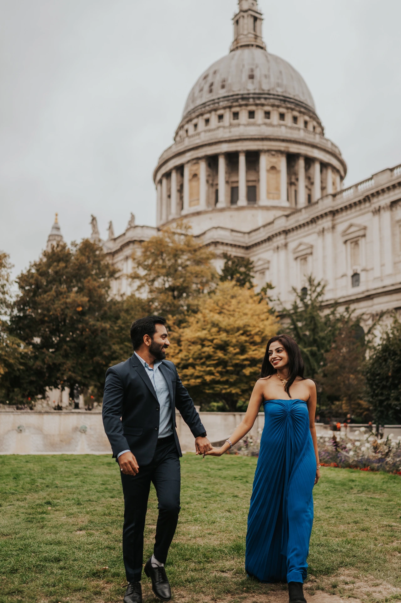 The couple holding hands on the Festival Gardens lawn, with their blue outfits against the cathedral and trees during their London proposal photoshoot.