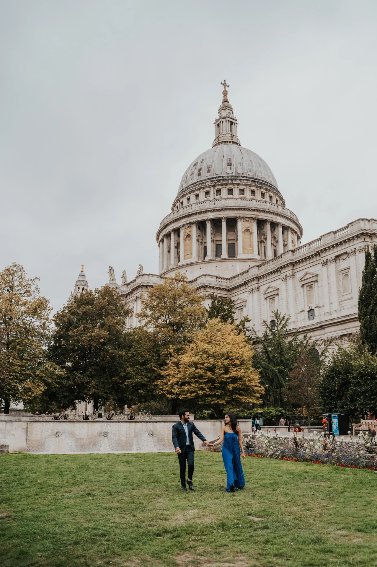 The couple holding hands on the Festival Gardens lawn beneath the dome of St Paul’s Cathedral during their London proposal photoshoot.