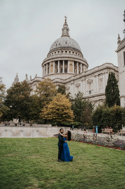 London proposal photoshoot: an embrace beneath St Paul’s dome The couple embracing on the Festival Gardens lawn, surrounded by trees beneath St Paul’s Cathedral during their London proposal photoshoot.
