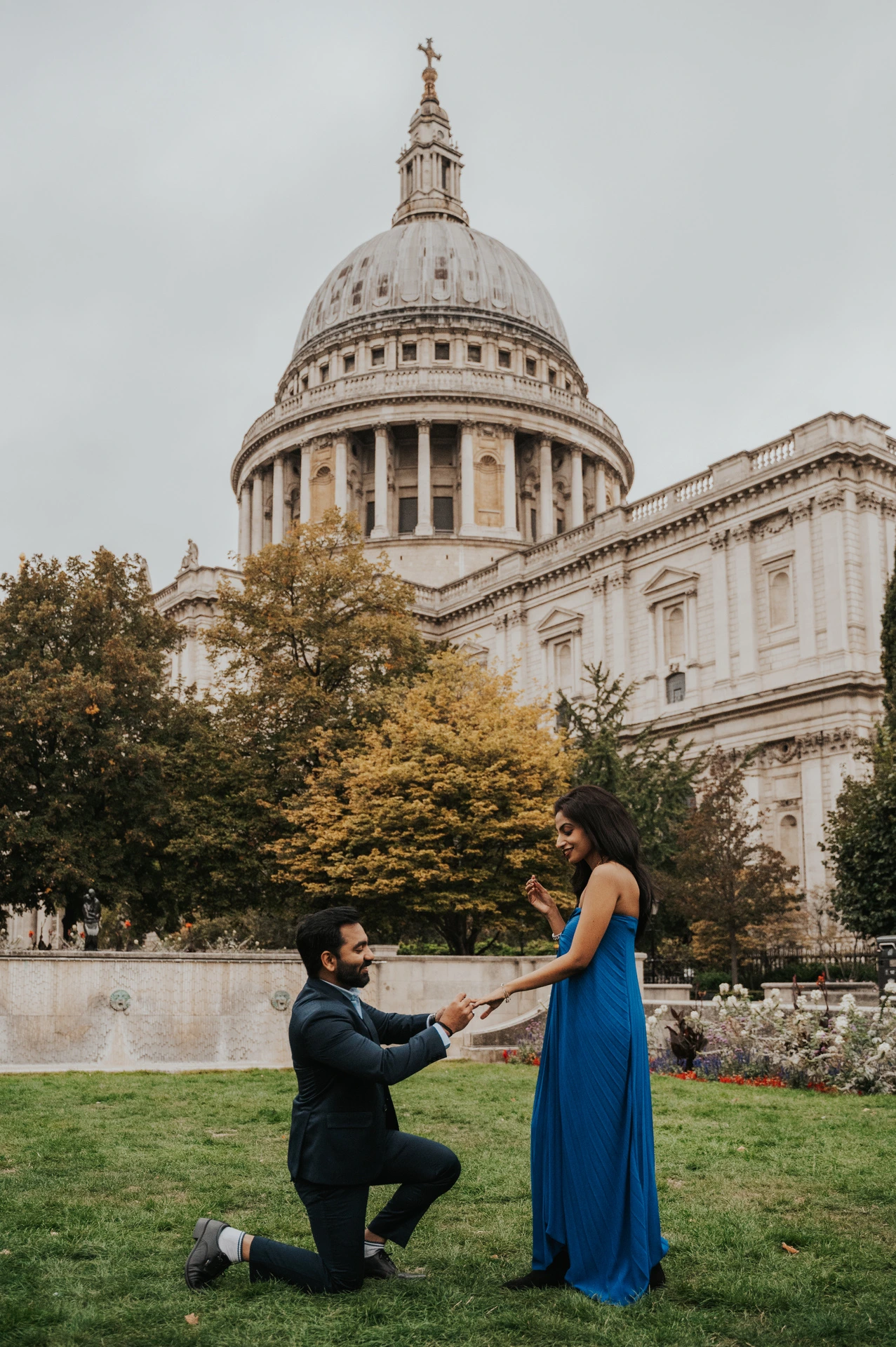 A man kneeling and holding his partner’s hand on the Festival Gardens lawn beneath St Paul’s Cathedral during their London proposal photoshoot.