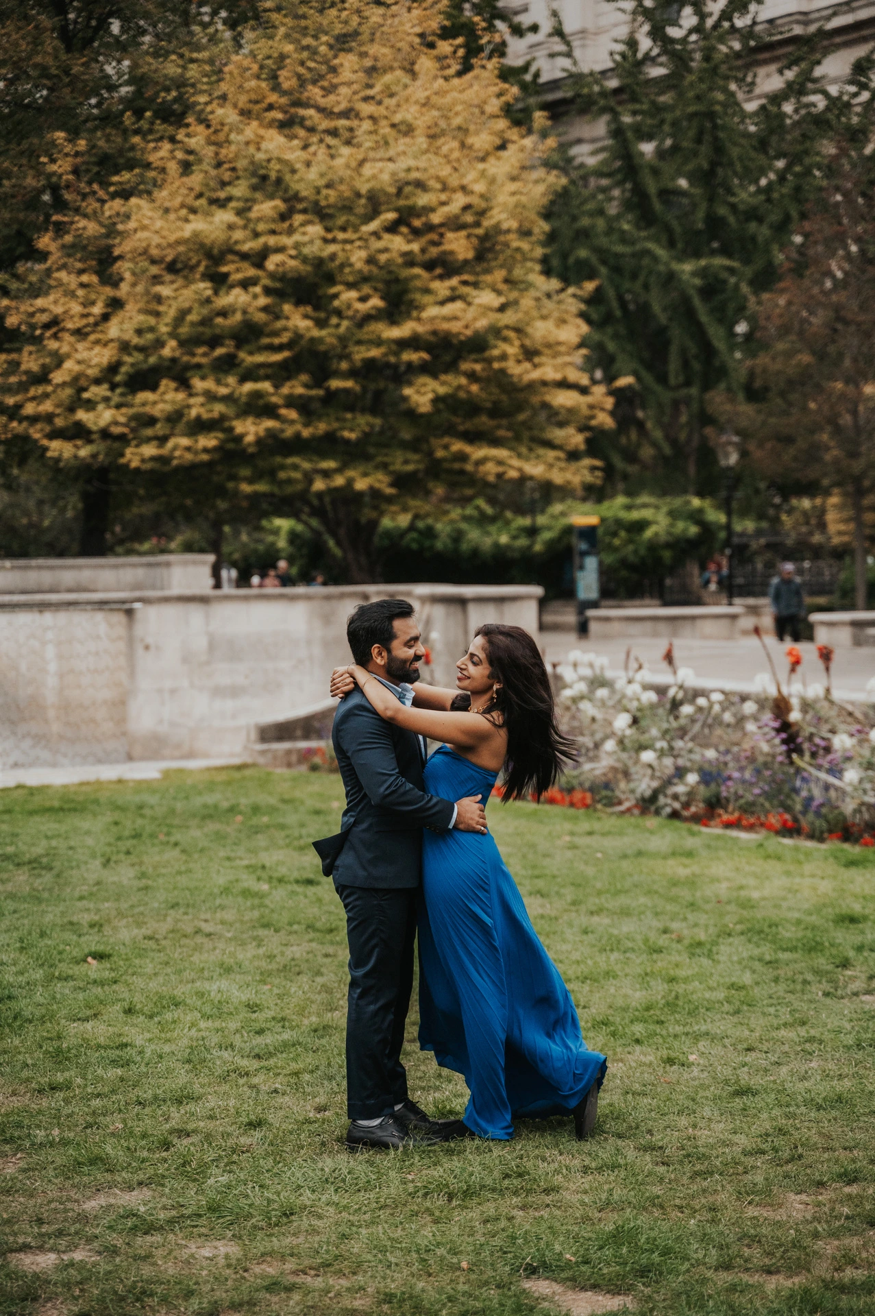 The couple smiling together on the Festival Gardens lawn as the woman lifts one foot in her blue dress during their London proposal photoshoot.