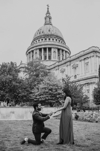 London proposal photoshoot: the proposal in black and white Black-and-white photograph of a man on one knee with his partner on the Festival Gardens lawn, framed by St Paul’s Cathedral during their London proposal photoshoot.