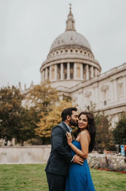 London proposal photoshoot: a smiling portrait in Festival Gardens The woman smiling as her partner kisses her cheek, with her blue dress and St Paul’s Cathedral in the view during their London proposal photoshoot.