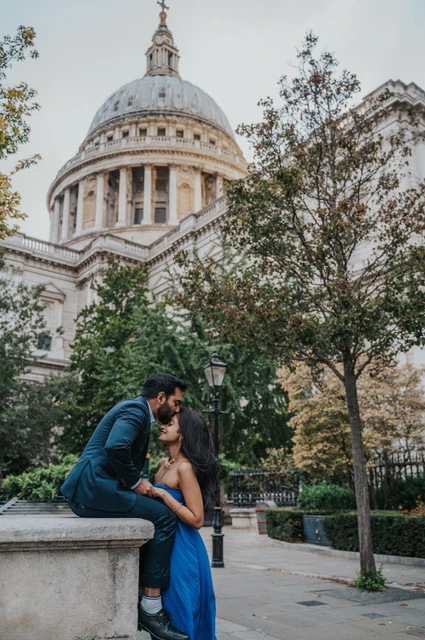 London proposal photoshoot: the garden’s stone wall The man seated on a stone wall as his partner stands beside him, with St Paul’s Cathedral above the trees during their London proposal photoshoot.