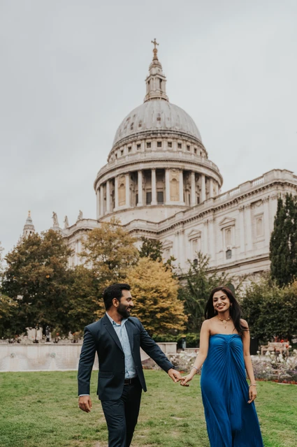London proposal photoshoot: walking in Festival Gardens The couple walking hand in hand on the lawn, her blue dress beside his dark suit, with St Paul’s Cathedral behind them during their London proposal photoshoot.