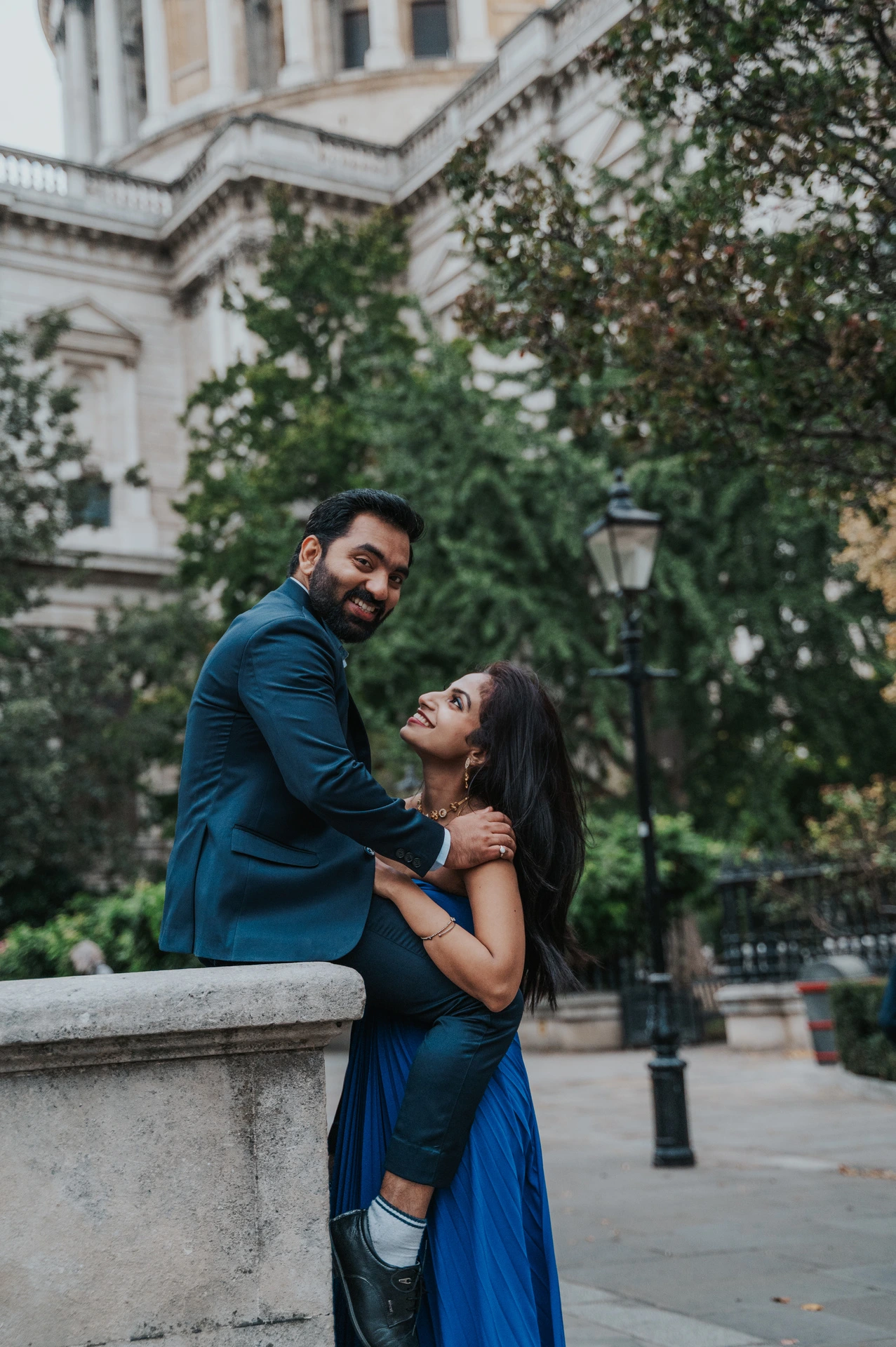 A closer portrait of the smiling couple beside a stone wall and garden trees near St Paul’s Cathedral during their London proposal photoshoot.