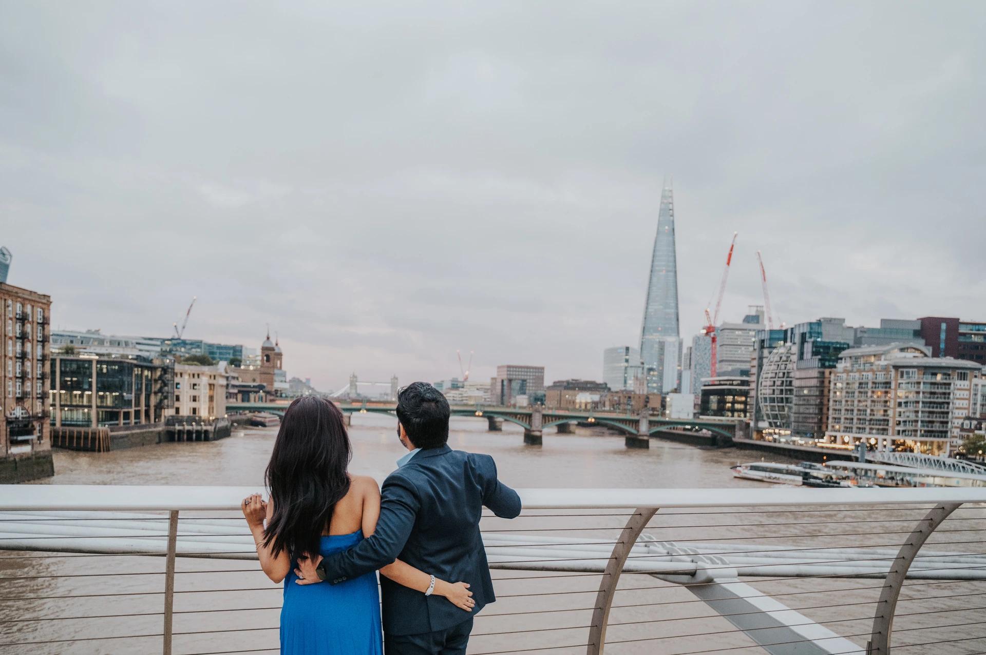 The couple looking across the Thames from Millennium Bridge towards the Shard and riverside buildings during their London proposal photoshoot.
