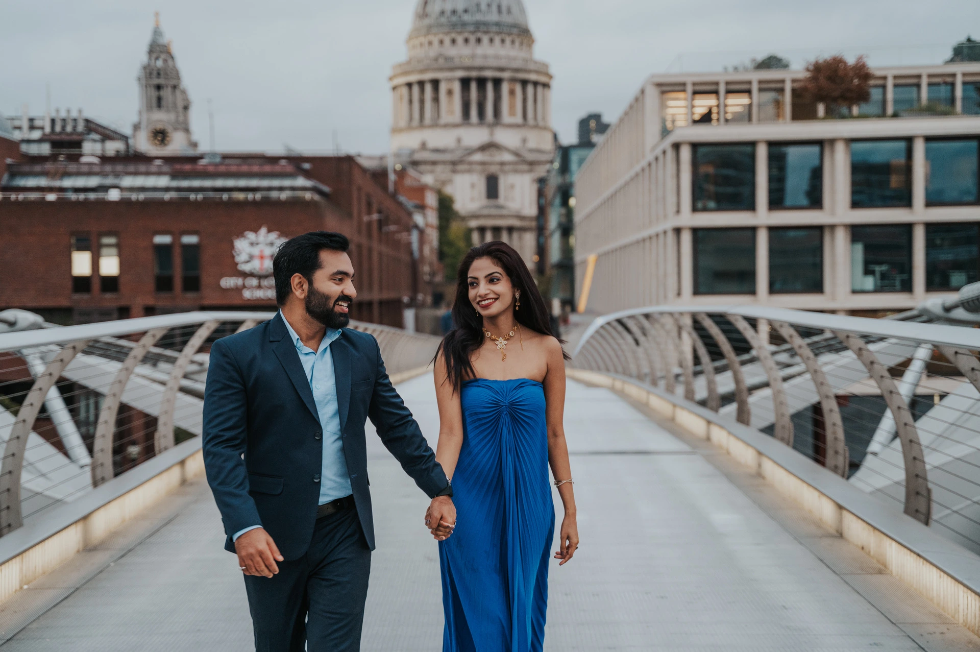A wide view of the couple walking along Millennium Bridge, with her blue dress and St Paul’s Cathedral behind them during their London proposal photoshoot.