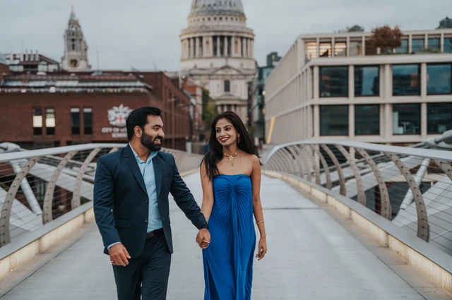 A wide view of the couple walking along Millennium Bridge, with her blue dress and St Paul’s Cathedral behind them during their London proposal photoshoot.