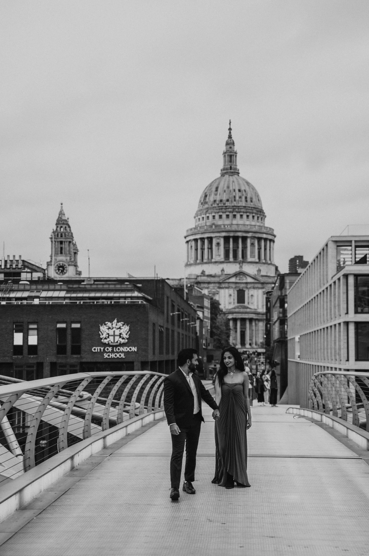 Black-and-white photograph of the couple walking together on Millennium Bridge beneath the skyline of St Paul’s Cathedral during their London proposal photoshoot.