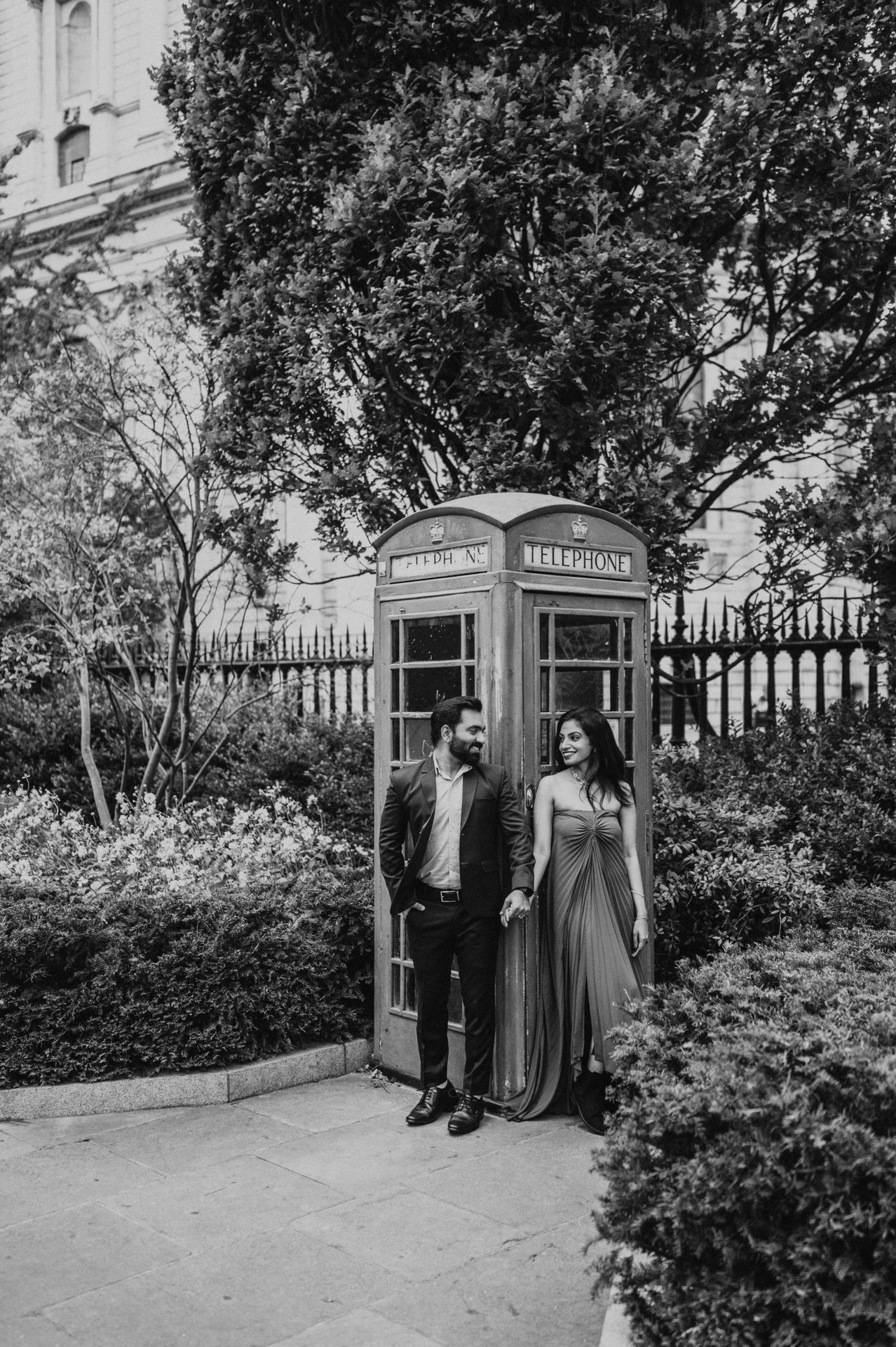 Black-and-white photograph of the couple together beside a telephone box beneath the trees near St Paul’s Cathedral during their London proposal photoshoot.