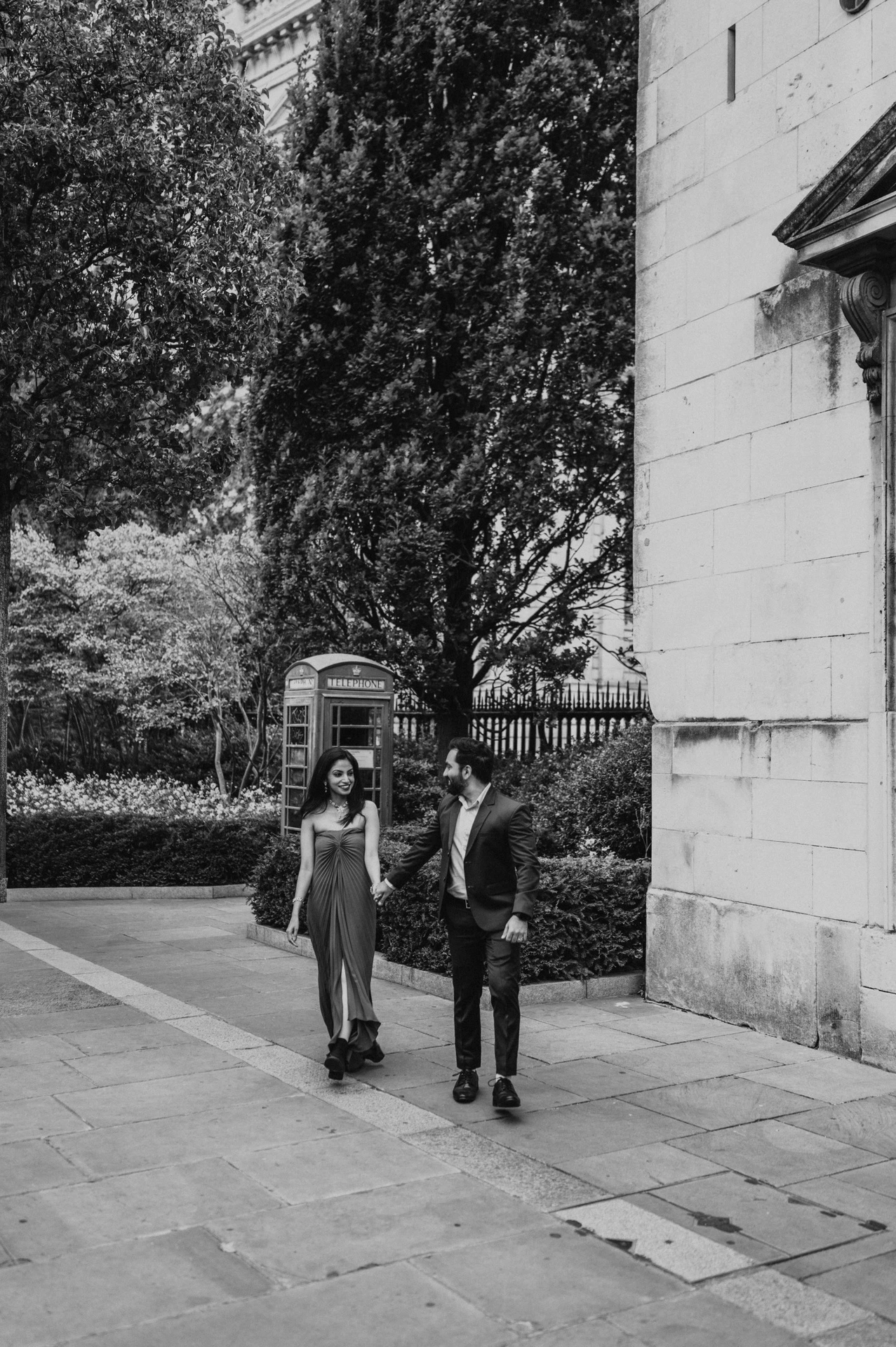 Black-and-white photograph of the couple walking beside a stone building and telephone box among the trees near St Paul’s Cathedral during their London proposal photoshoot.