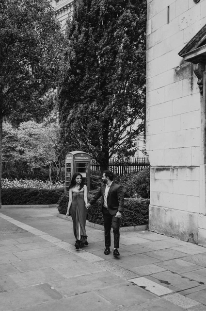 London proposal photoshoot: the telephone box walk in black and white Black-and-white photograph of the couple walking beside a stone building and telephone box among the trees near St Paul’s Cathedral during their London proposal photoshoot.
