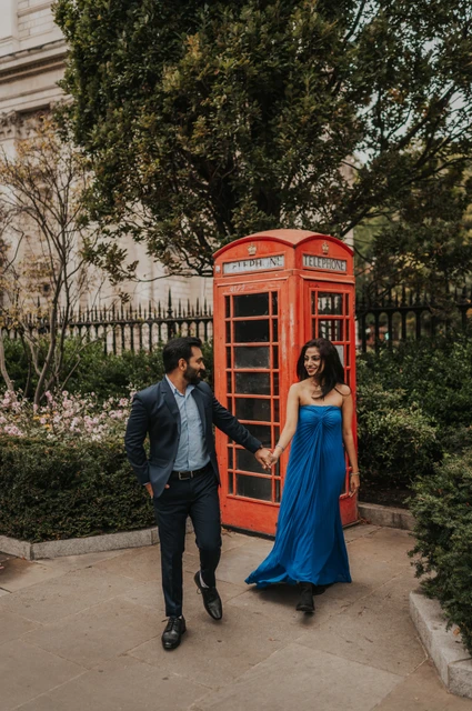 London proposal photoshoot: a stroll beside the red telephone box The couple holding hands as they walk away from a red telephone box beside garden railings during their London proposal photoshoot.