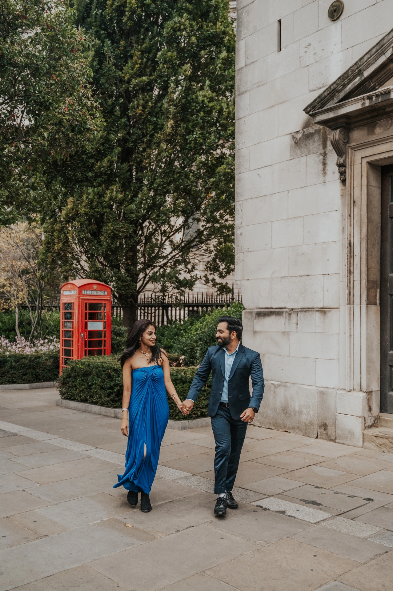 The couple walking hand in hand beside a pale stone building and a red telephone box near St Paul’s Cathedral during their London proposal photoshoot.