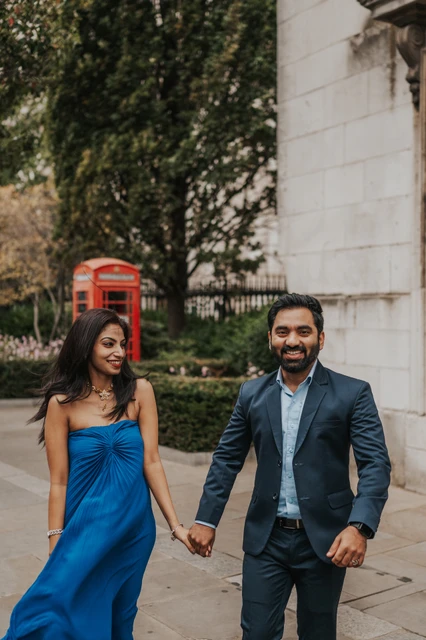 London proposal photoshoot: smiles beside the red telephone box The smiling couple walking towards the camera, with a red telephone box and trees behind them during their London proposal photoshoot.