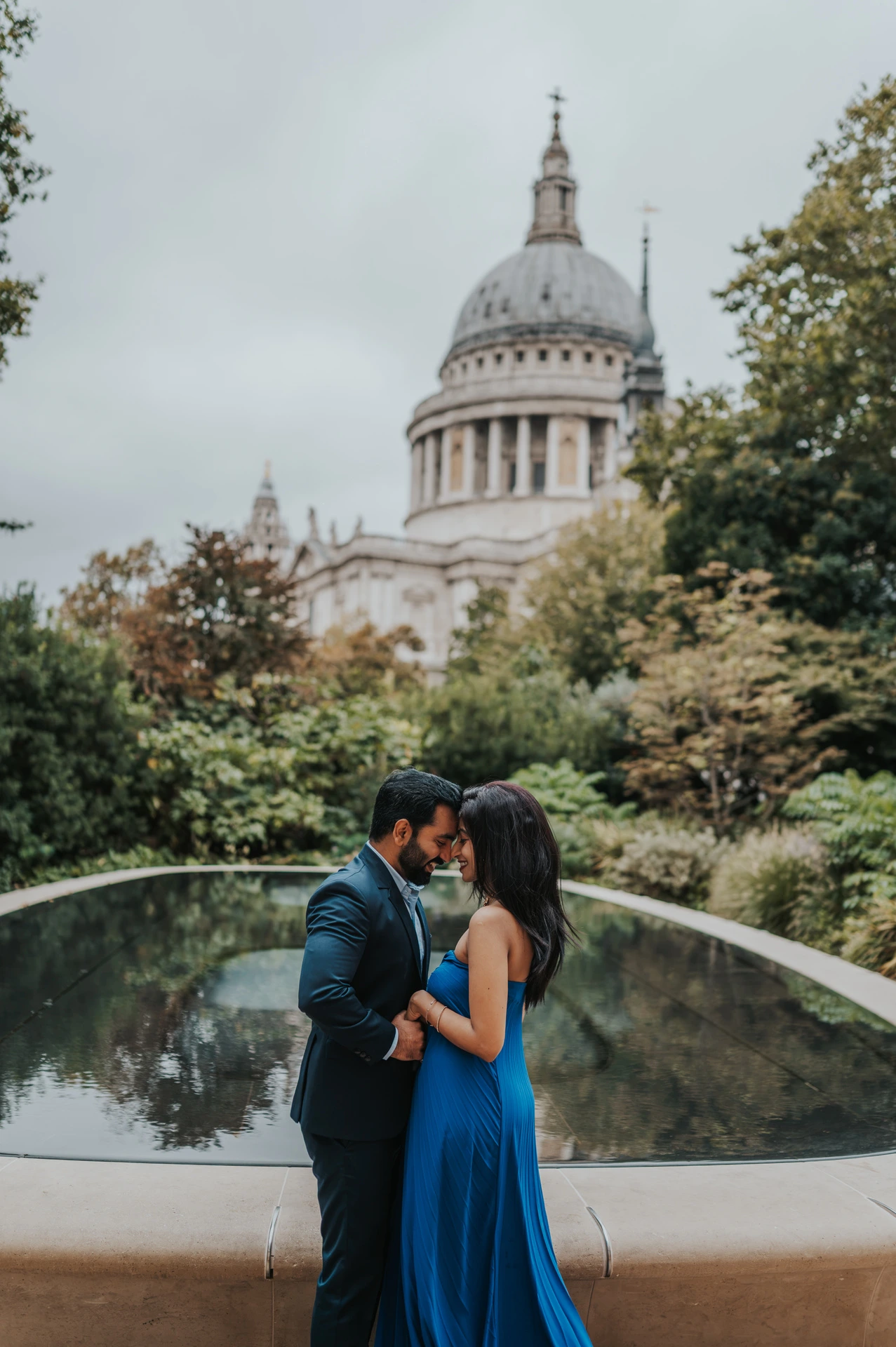 The couple leaning close together beside the Reflection Garden pool, framed by trees and St Paul’s Cathedral during their London proposal photoshoot.