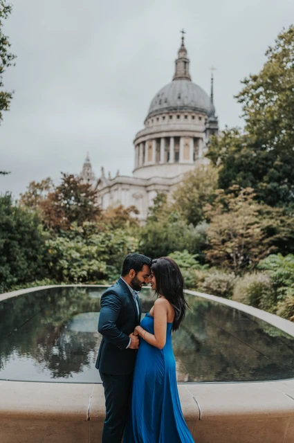 London proposal photoshoot: a close portrait beside the reflecting pool The couple leaning close together beside the Reflection Garden pool, framed by trees and St Paul’s Cathedral during their London proposal photoshoot.