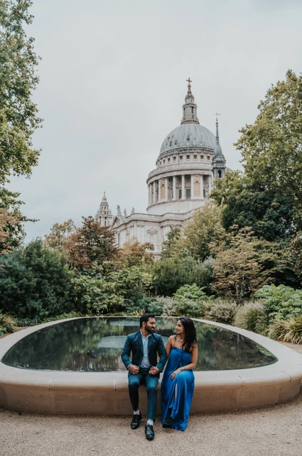 London proposal photoshoot: seated beside the Reflection Garden pool The couple sitting on the curved edge of the Reflection Garden pool beneath a view of St Paul’s Cathedral during their London proposal photoshoot.