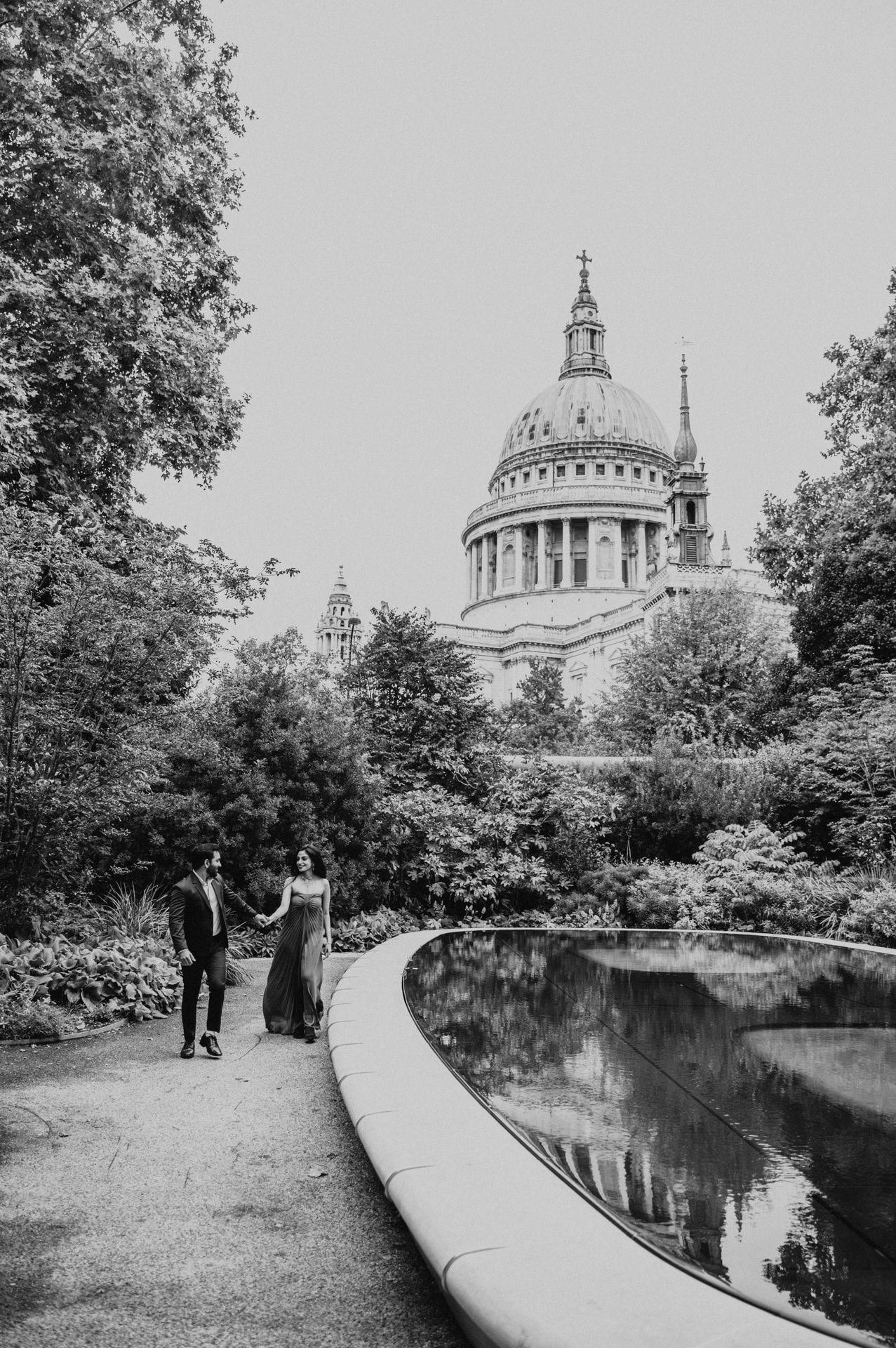 Black-and-white photograph of the couple holding hands beside the curved reflecting pool and leafy planting with St Paul’s Cathedral above during their London proposal photoshoot.