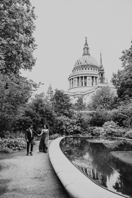 London proposal photoshoot: the Reflection Garden in black and white Black-and-white photograph of the couple holding hands beside the curved reflecting pool and leafy planting with St Paul’s Cathedral above during their London proposal photoshoot.