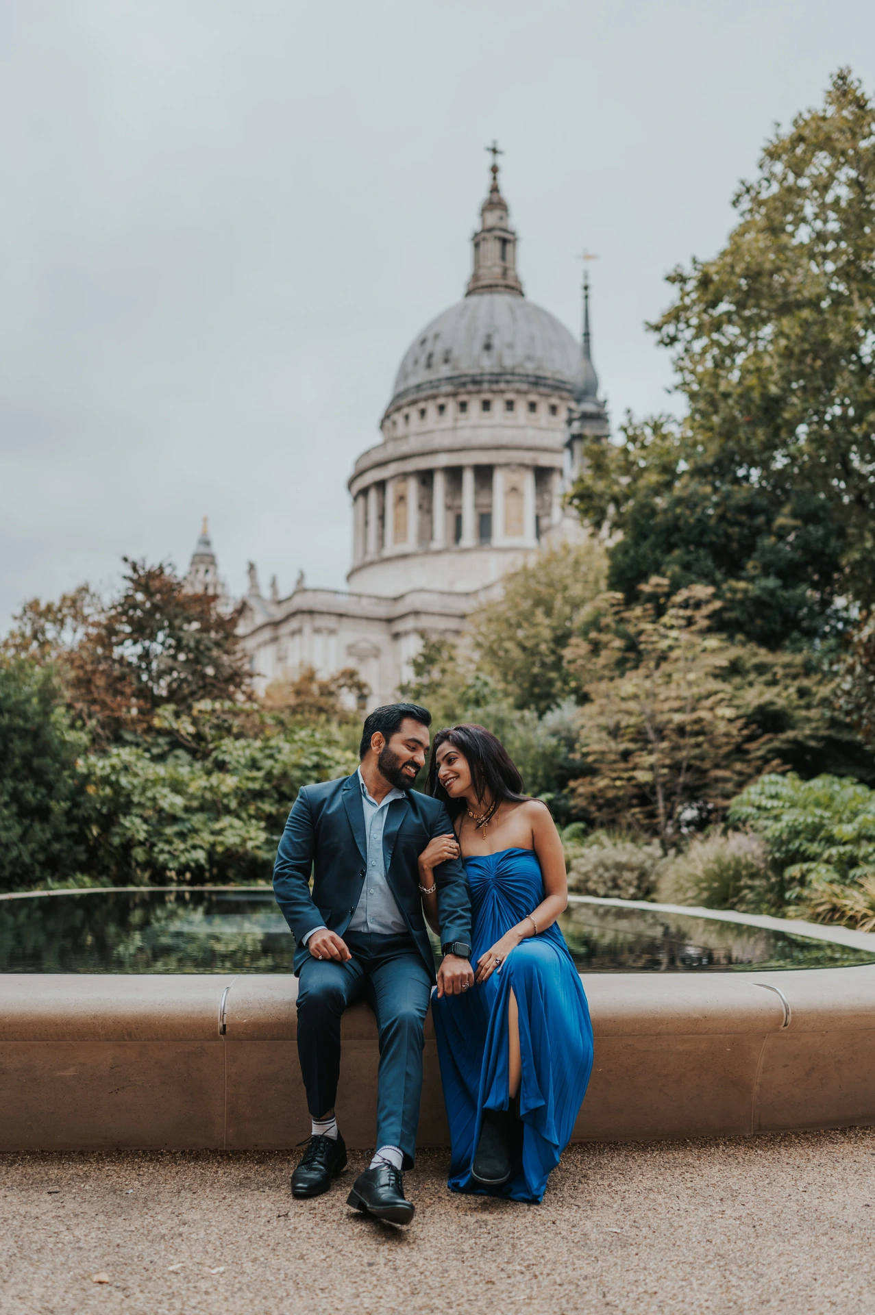 The couple leaning close together while sitting beside the Reflection Garden pool, with the cathedral dome above the trees during their London proposal photoshoot.