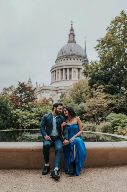 The couple leaning close together while sitting beside the Reflection Garden pool, with the cathedral dome above the trees during their London proposal photoshoot.