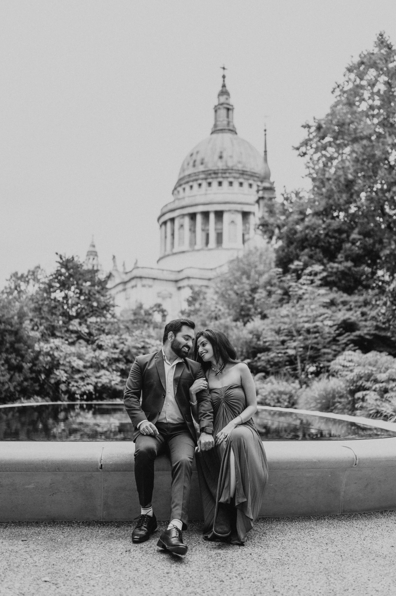 Black-and-white photograph of the couple sitting close together beside the Reflection Garden pool beneath St Paul’s Cathedral during their London proposal photoshoot.