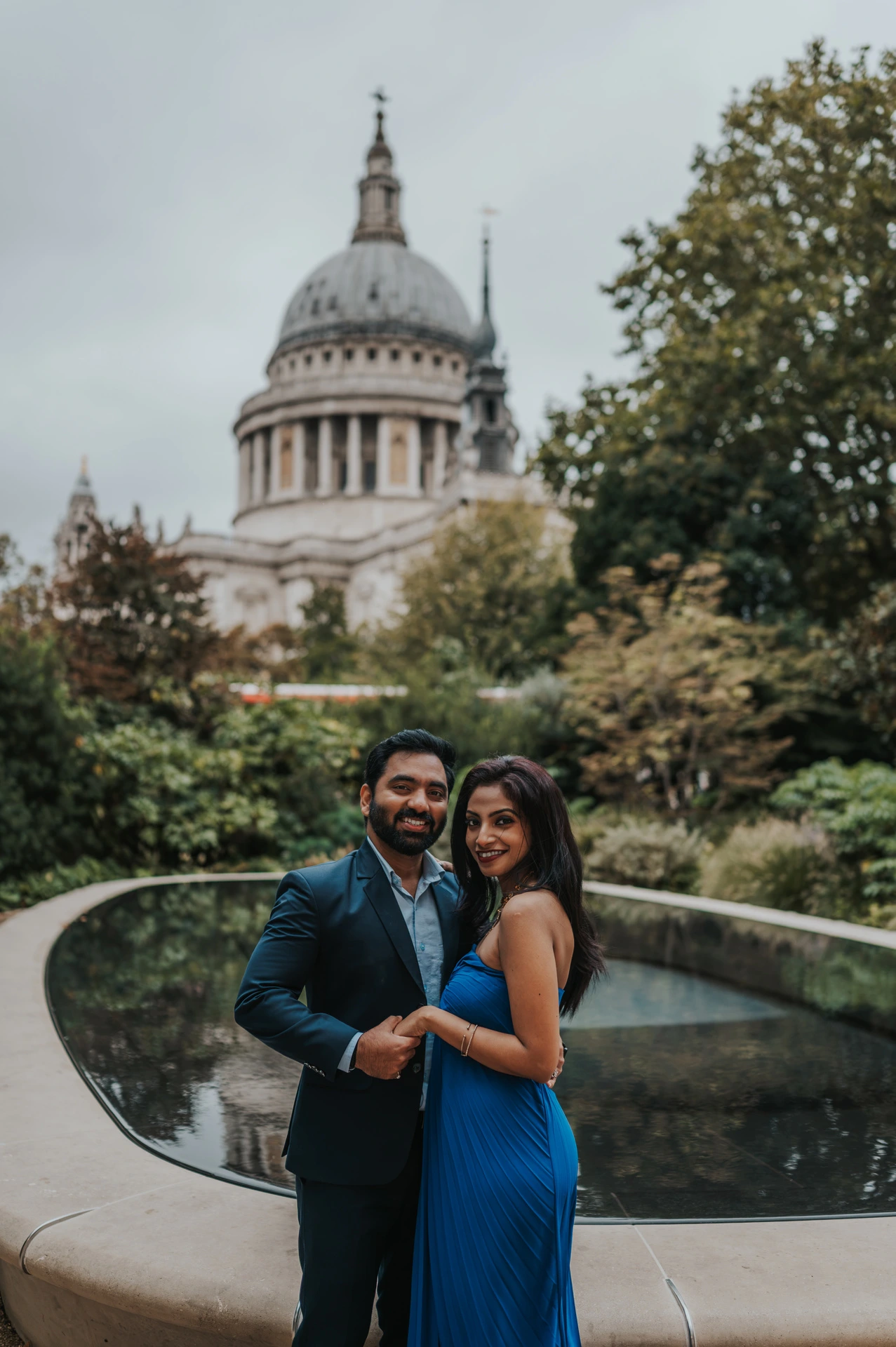 The couple smiling together by the Reflection Garden pool with St Paul’s Cathedral behind them during their London proposal photoshoot.