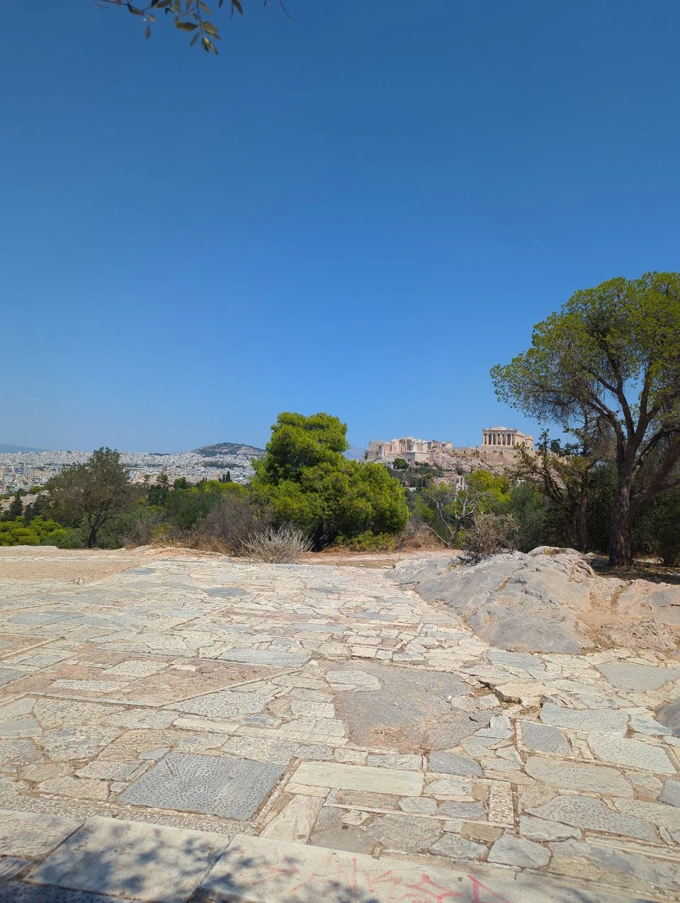Acropolis Photography Alternatives - Philopappos View A paved viewpoint on lower Philopappos faces the Acropolis across trees and the Athens skyline under a clear blue sky.