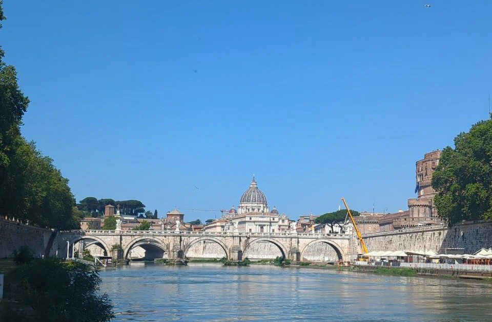Rome Photoshoot Locations - Lower Tiber View The Tiber flows beneath a stone bridge toward St Peter's dome, with Castel Sant'Angelo rising at the right in Rome.