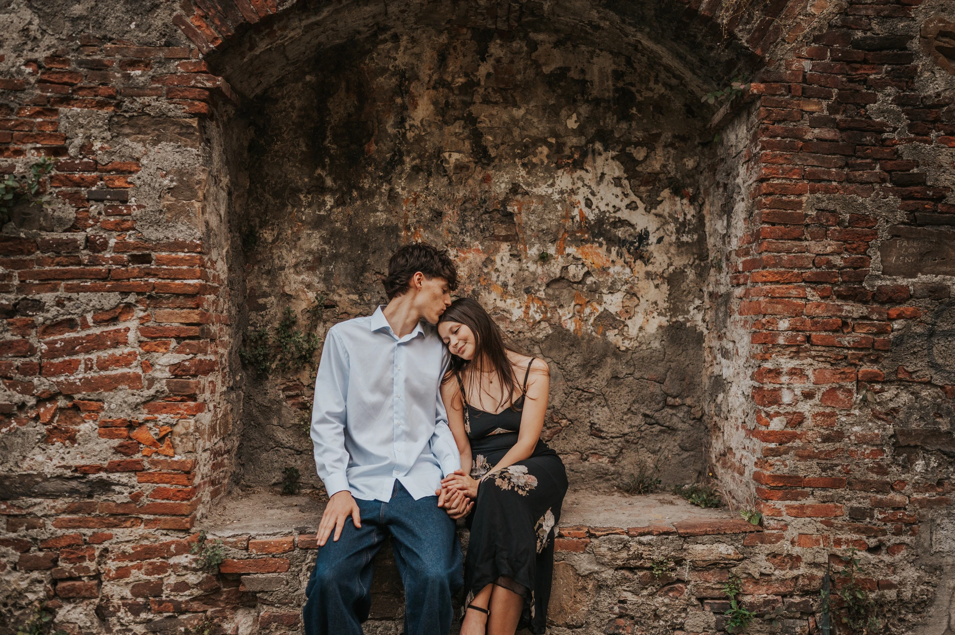 Alice and Pablo sit together in a brick alcove, her floral dress contrasting with his pale blue shirt. From Alice and Pablo’s Lucca anniversary photoshoot.