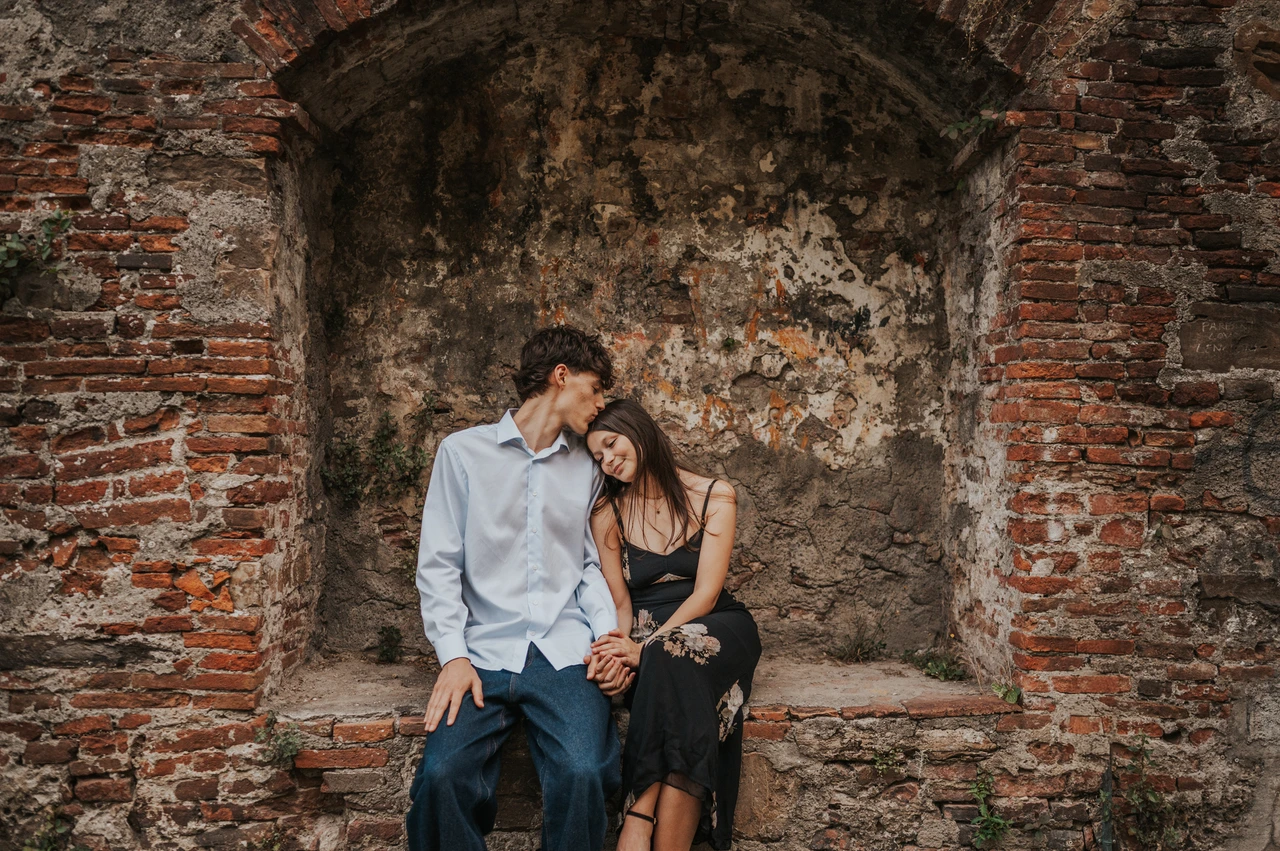 A seated couple portrait during a Lucca anniversary photoshoot Alice and Pablo sit together in a brick alcove, her floral dress contrasting with his pale blue shirt. From Alice and Pablo’s Lucca anniversary photoshoot.