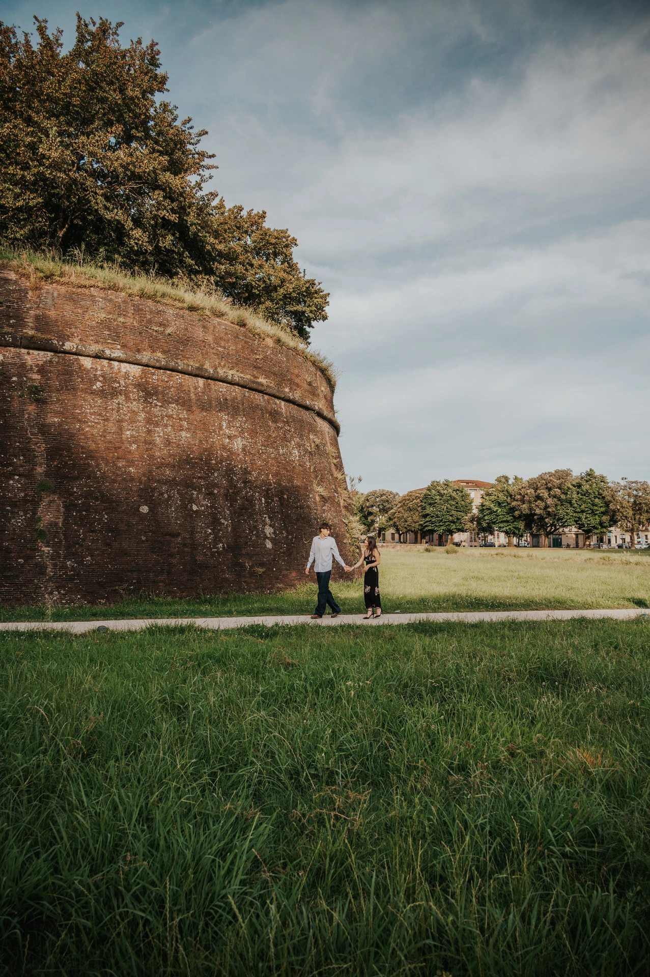 Alice and Pablo hold hands on the green lawn below Lucca’s curved brick walls. From Alice and Pablo’s Lucca anniversary photoshoot.