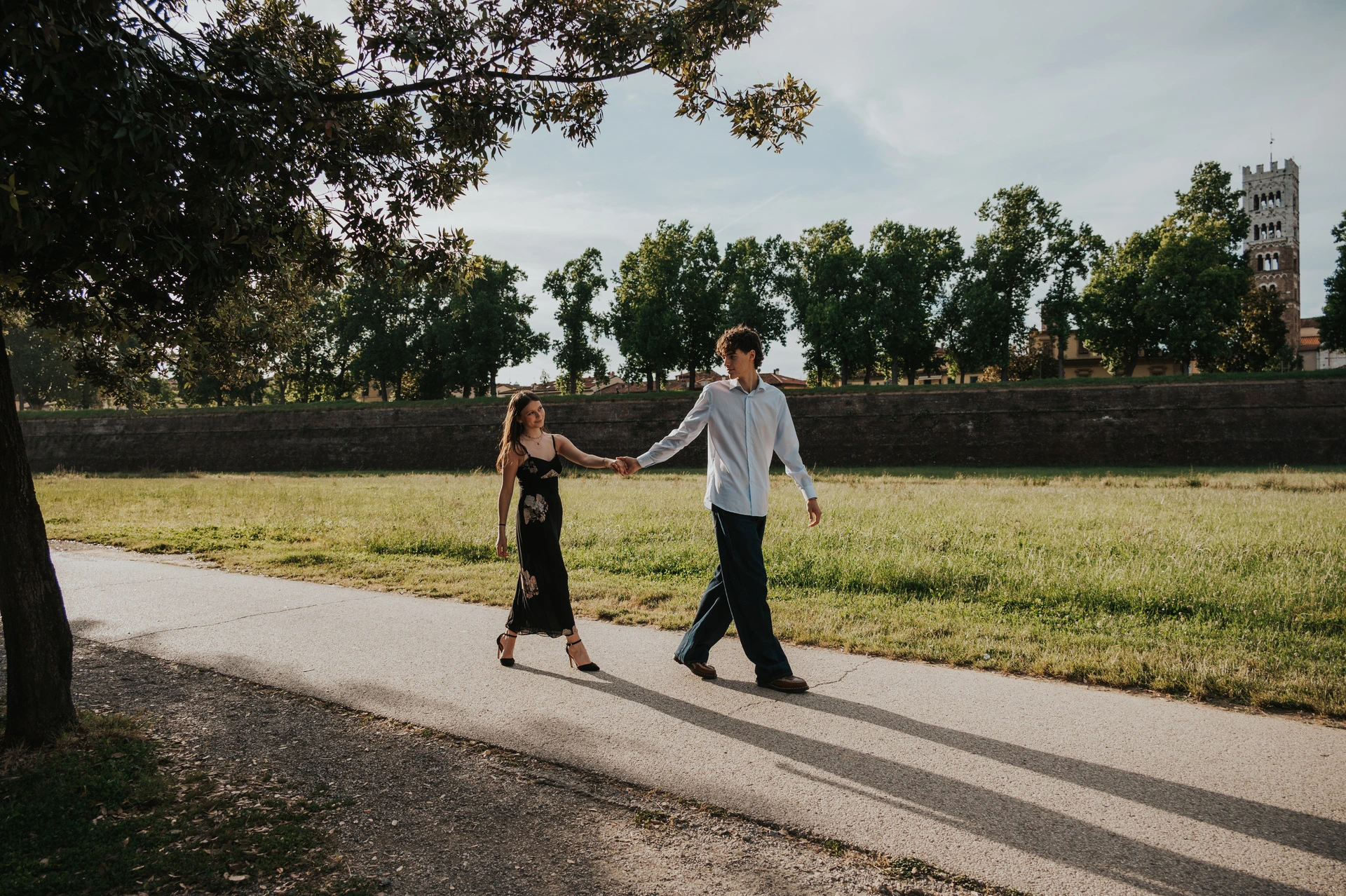 Alice and Pablo walk along the path on Lucca’s city walls, holding hands beside the grass. From Alice and Pablo’s Lucca anniversary photoshoot.