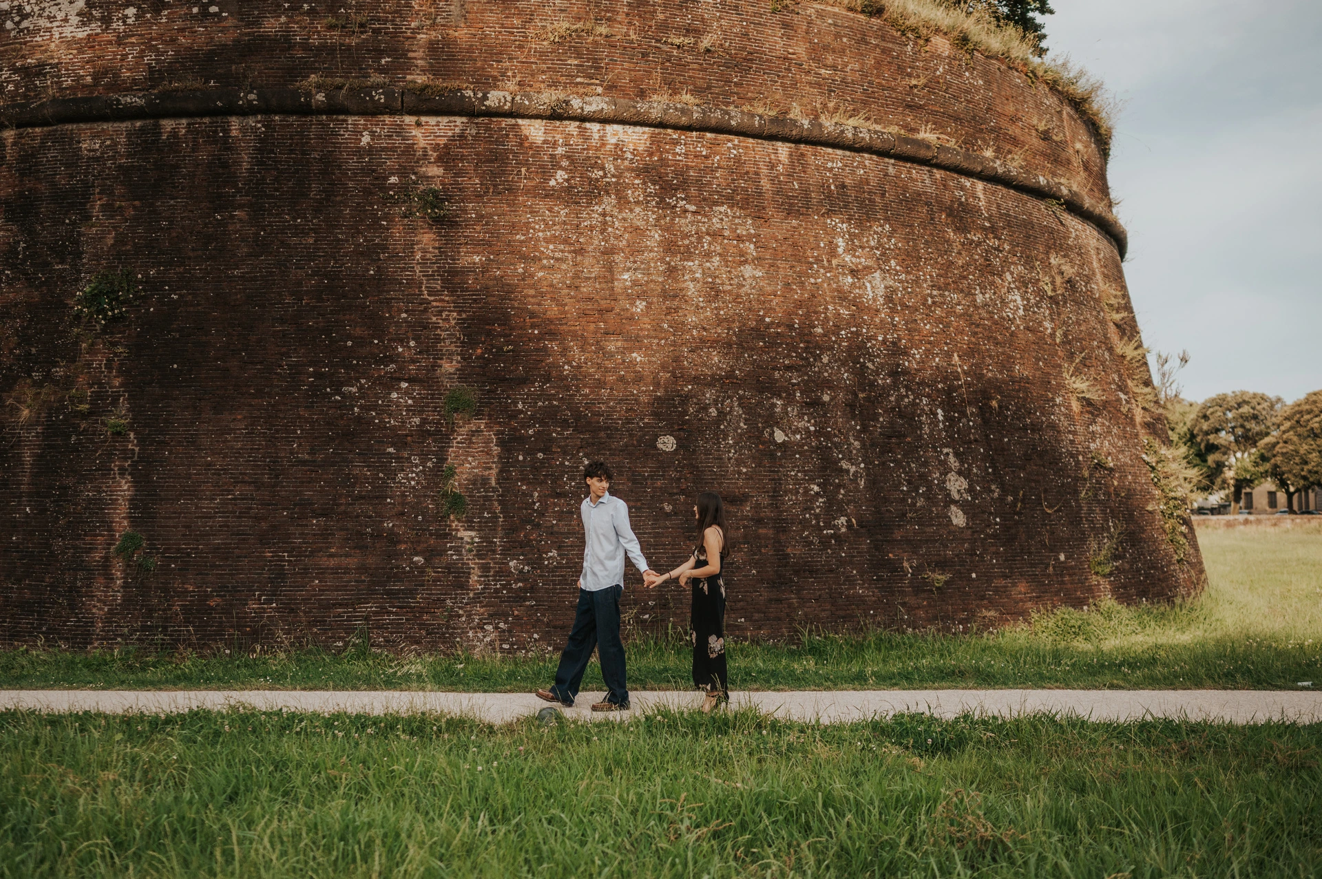 Alice and Pablo walk hand in hand beside the tall brick city walls. From Alice and Pablo’s Lucca anniversary photoshoot.