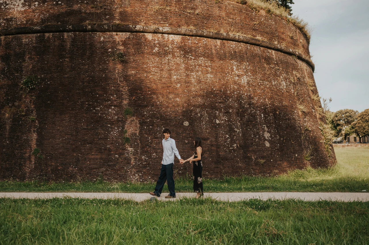 Walking by the city walls during a Lucca couple photoshoot Alice and Pablo walk hand in hand beside the tall brick city walls. From Alice and Pablo’s Lucca anniversary photoshoot.