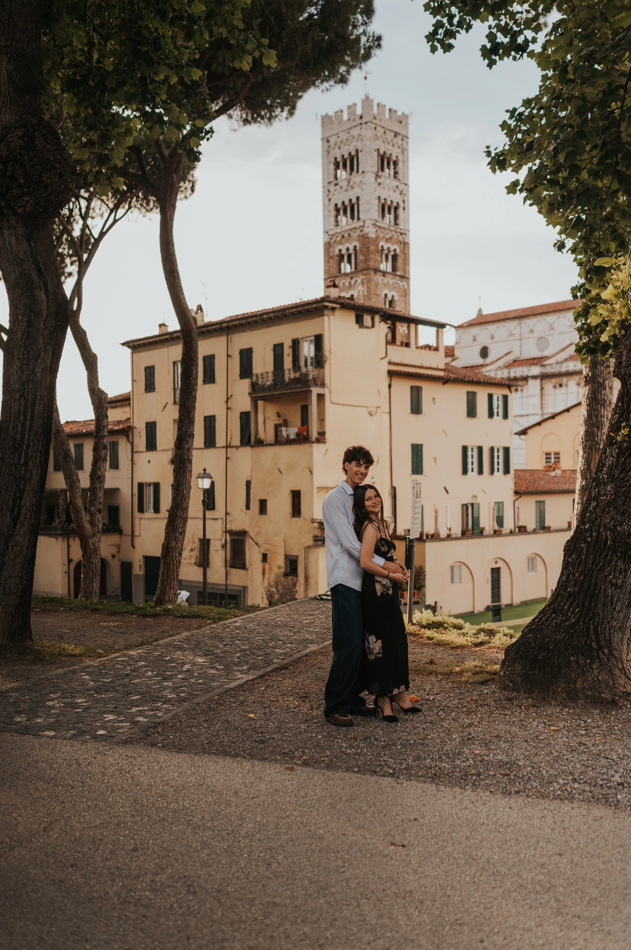 Alice and Pablo stand beneath tall trees with Lucca’s Duomo and bell tower behind them. From Alice and Pablo’s Lucca anniversary photoshoot.