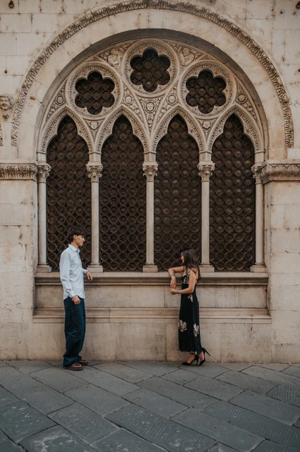 Lucca couple photoshoot by an ornate Gothic window Alice and Pablo stand apart holding hands in front of an ornate Gothic window in Lucca’s old town. From Alice and Pablo’s Lucca anniversary photoshoot.