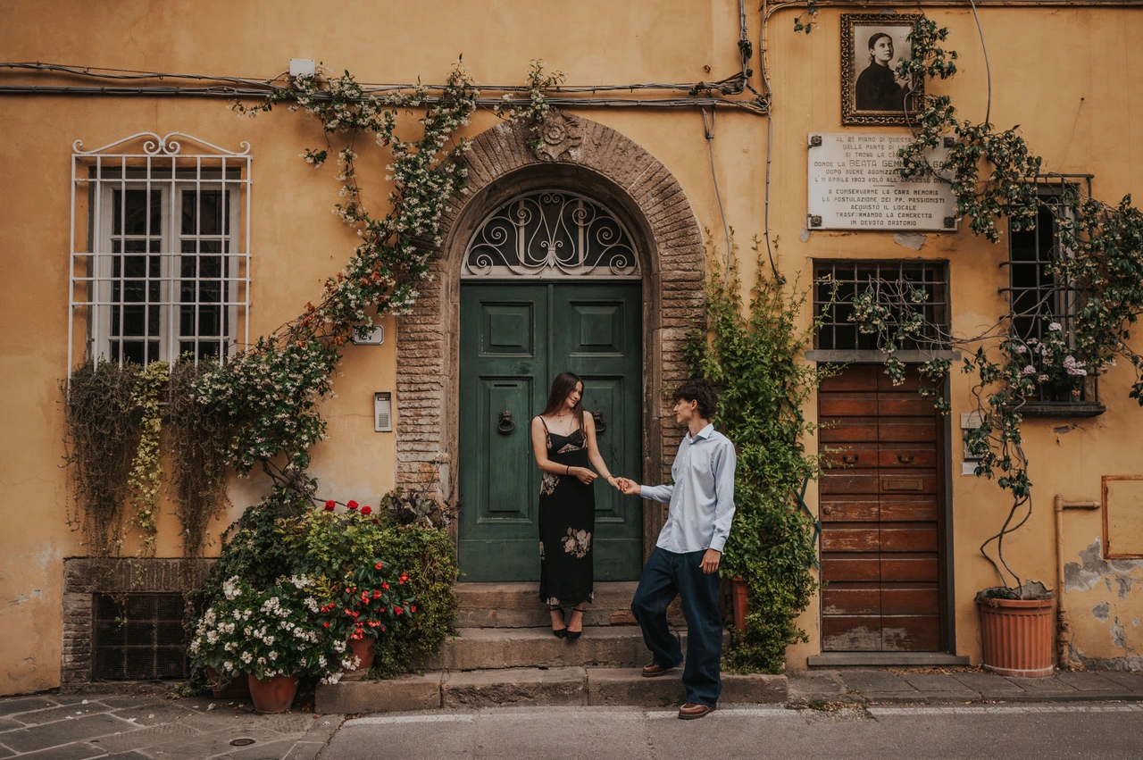 Warm old-town colours in a Lucca couple photoshoot Alice and Pablo stand by a green door beneath climbing flowers, with ochre walls and shuttered windows around them. From Alice and Pablo’s Lucca anniversary photoshoot.